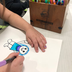 photograph of child drawing, only the arms and hands of the child are in the picture, one hand is holding a marker, drawing is child wearing blue with blue hair, a box of markers in the photo as well